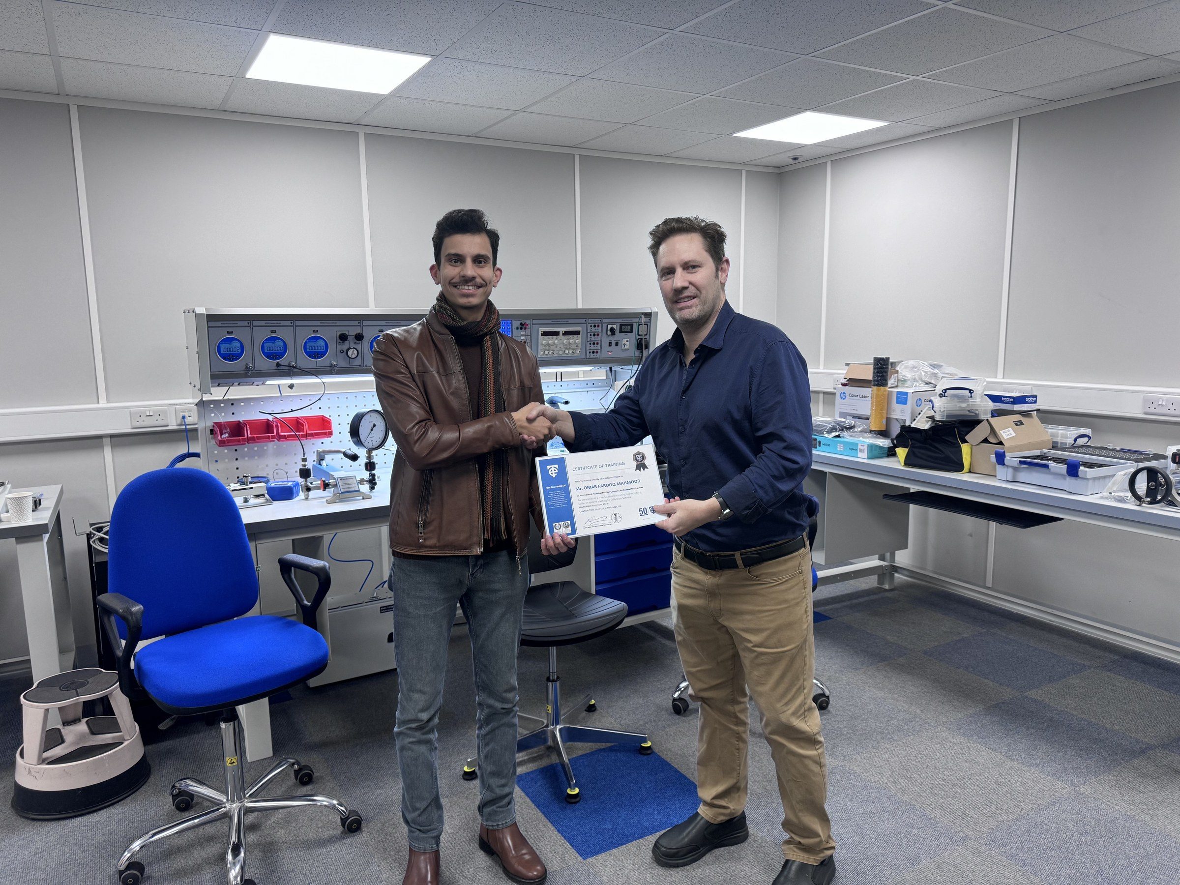 Omar Farooq Al Obaidi and Robert Bailey shaking hands over Omar's Certificate of Training in front of the calibration bench.